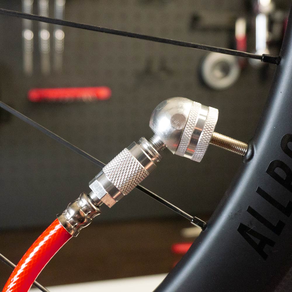 A close-up of a bicycle tire valve being inflated using a red air hose and the Prestacycle Big Angle Presta Head. The blurred workshop background features tools on the wall and tubeless valves ready for use.