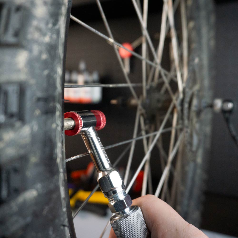 A hand uses the Prestaflator Mini Pro – Presta / Schrader push-to-inflate Bicycle Inflation Tool to check a bike tire’s air pressure, with the gauge on the valve and bike spokes visible in the background.