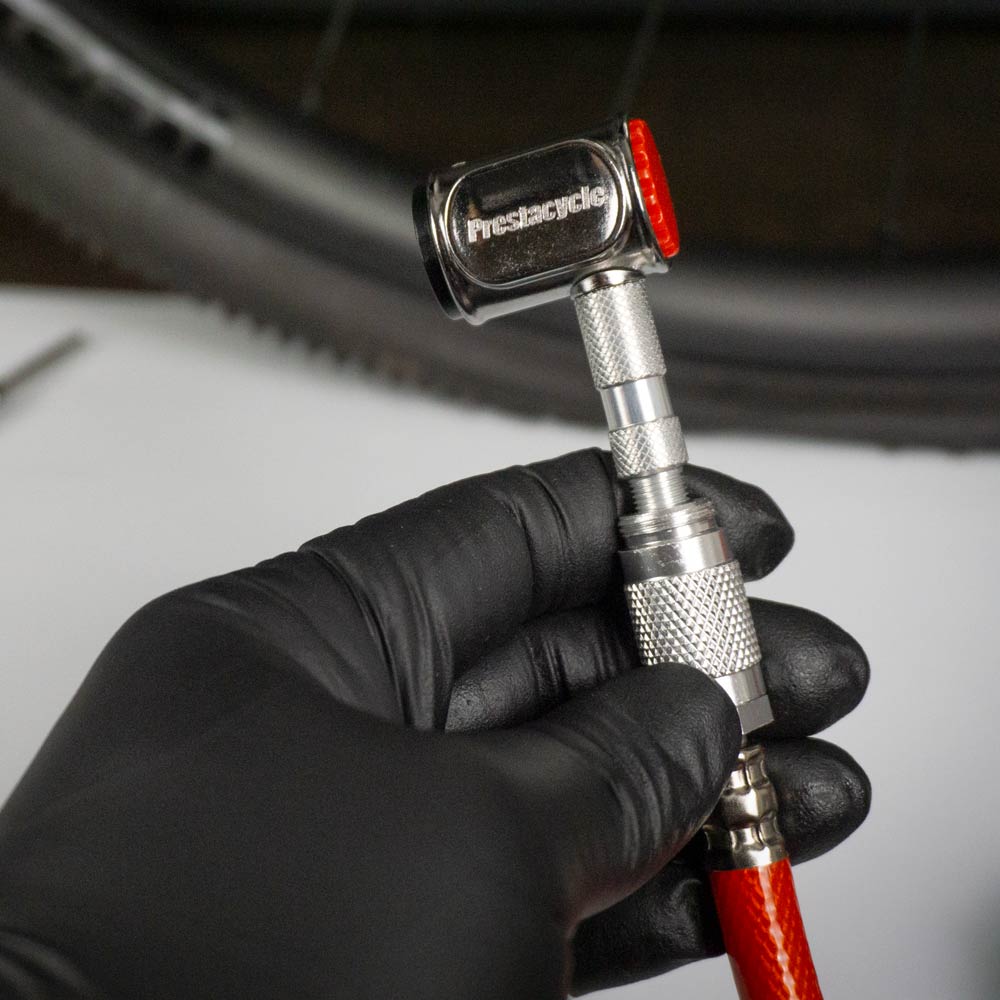 A gloved hand holds a Prestaflator PRO Digital Bicycle Tyre Inflator, featuring machined brass internals and attached to a red hose, with a bicycle tire in the background.
