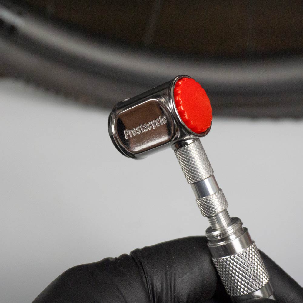 A gloved hand holds the Prestacycle Pro Presta Head, a silver tool with a red cap and auto-lock feature, positioned near a bicycle tire valve.