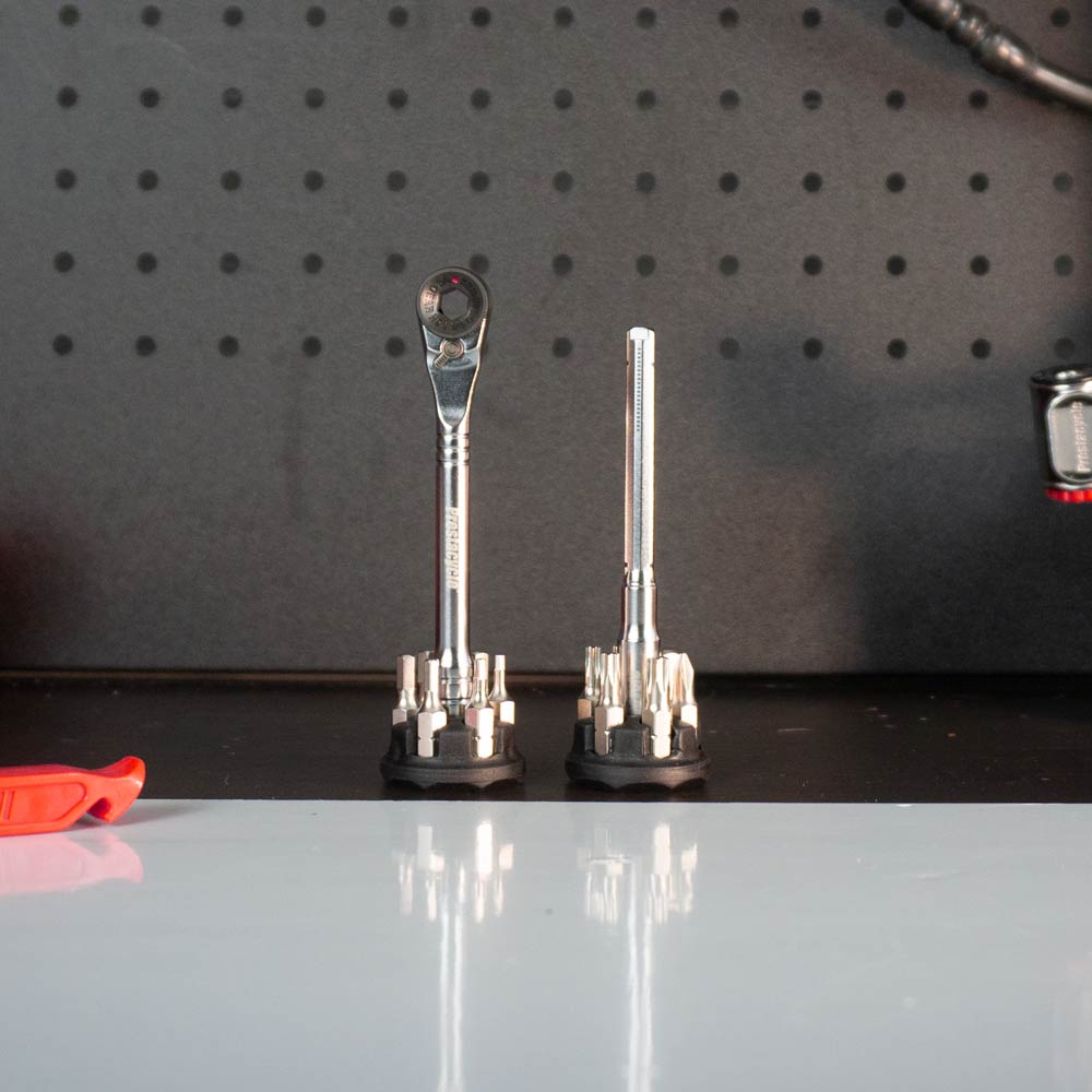 The Prestacycle Pro T-Handle Kit with 12 S2 bits and 2 round holders stands upright on a black workbench before a pegboard wall, with an orange tool partially visible on the left.