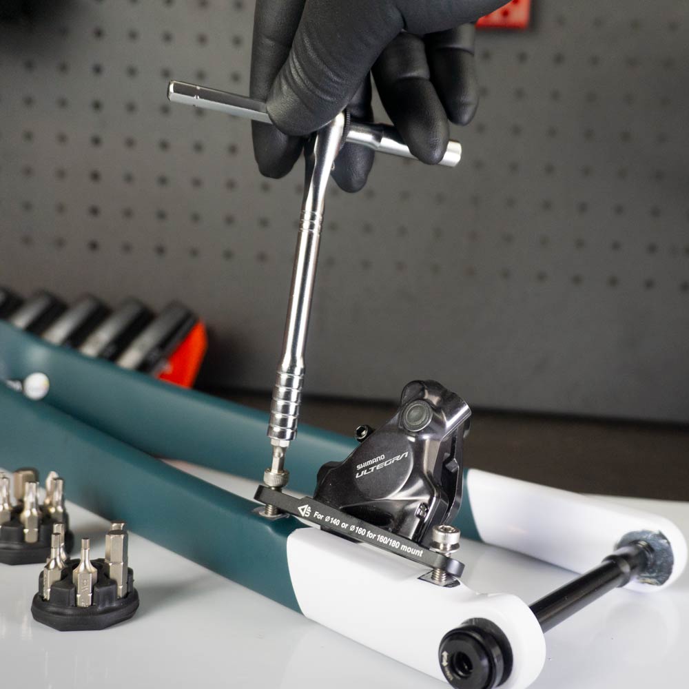 A person wearing a black glove uses the Prestacycle Pro T-Handle - Kit with (12) S2 and (2) Round Holder to adjust a bicycle disc brake caliper, with assorted tool bits, a ratchet set, and a pegboard in the background.