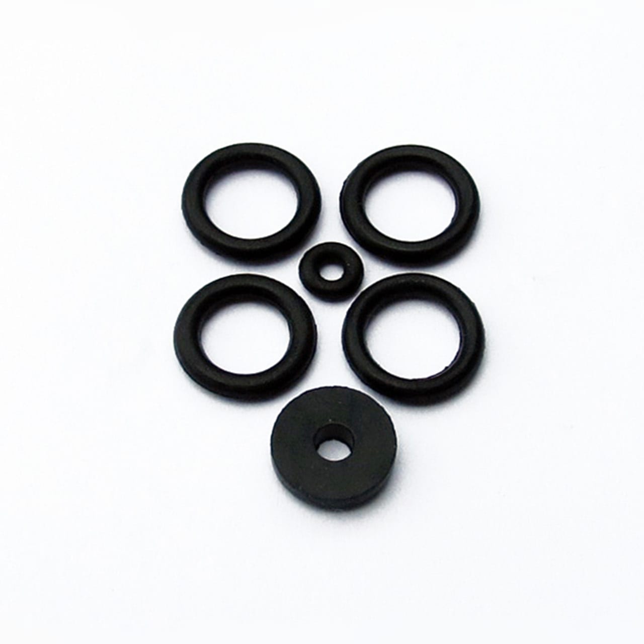 Five black o-rings and one solid black rubber washer from the Prestaflator Rebuild Kit are arranged on a white background to resemble a simple face with eyes, nose, mouth, and chin.