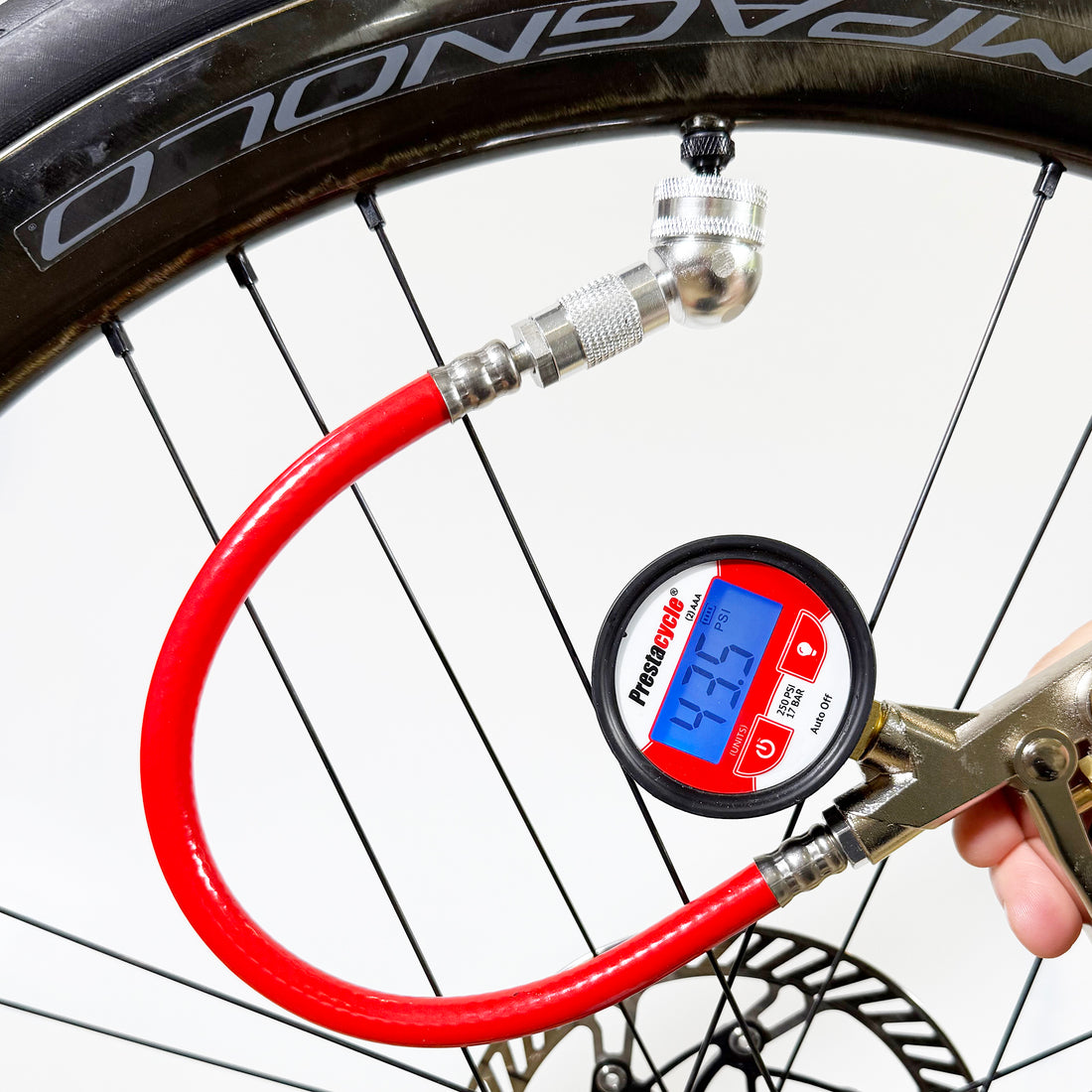 A close-up of a person using the Prestacycle Prestaflator Digital Bicycle Tire Inflator with a red hose to check bicycle tire pressure. The Presta &amp; Schrader air compressor tool displays a reading on its digital screen and is tubeless-safe.