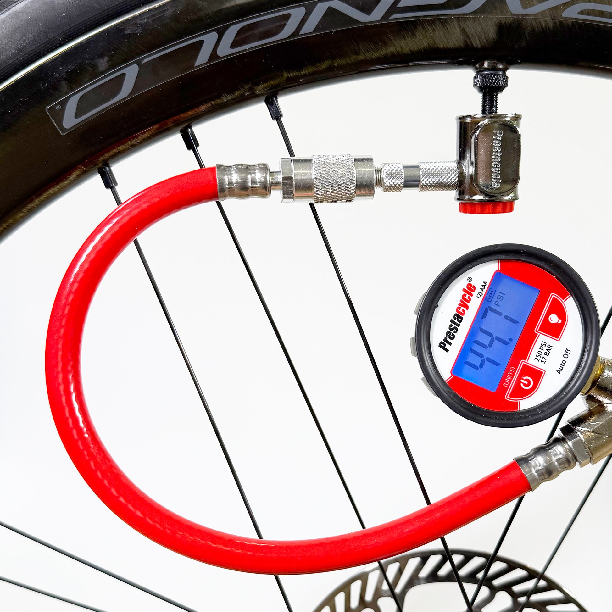 The Prestaflator PRO Digital Bicycle Tyre Inflator, with machined brass internals and a red hose, is attached to a bicycle Presta valve and displays 40.1 PSI on a white background; the black wheel and spokes are visible.
