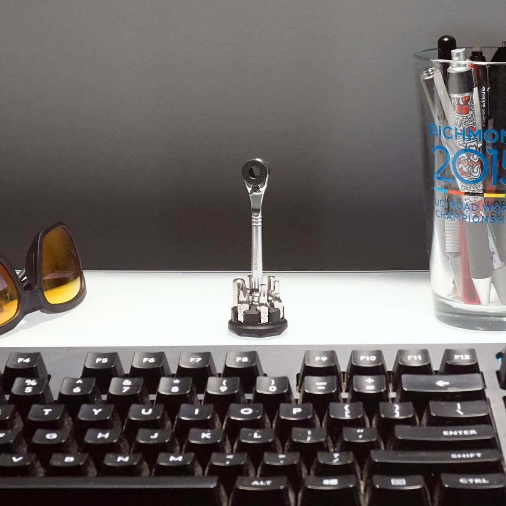 A close-up of a desk features a keyboard, sunglasses, pens in a glass, and the Prestaratchet Micro – Kit with 6 S2 bits and round holder stand on a white surface against a gray background.