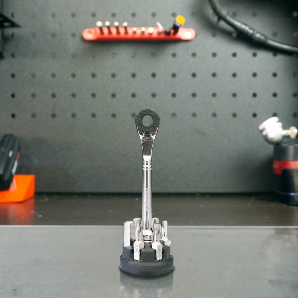 The Prestaratchet Micro – Kit with (6) S2 Bits and a Round Holder Stand is displayed upright on a workbench, while a pegboard and assorted tools hang in the blurred background.