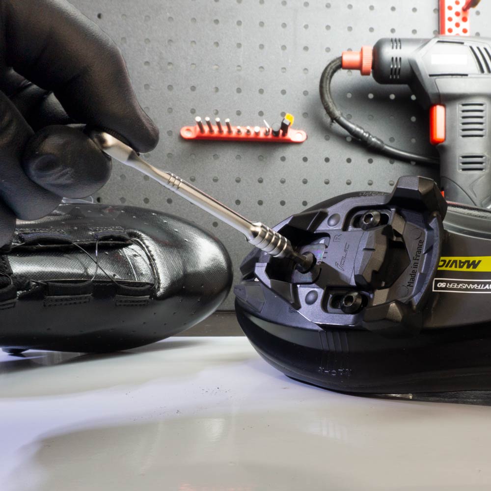 A hand in a black glove uses a screwdriver to install a cycling cleat onto the sole of a black cycling shoe, with the Prestaratchet Micro – Kit with 6 S2 Bits and Round Holder Stand visible in the background.
