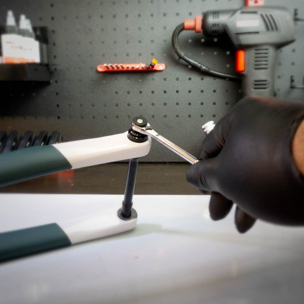 Wearing a black glove, a person uses the Prestaratchet Micro – Kit with (6) S2 Bits and Round Holder Stand to adjust a bolt on a bicycle fork in a workshop surrounded by tools and equipment.