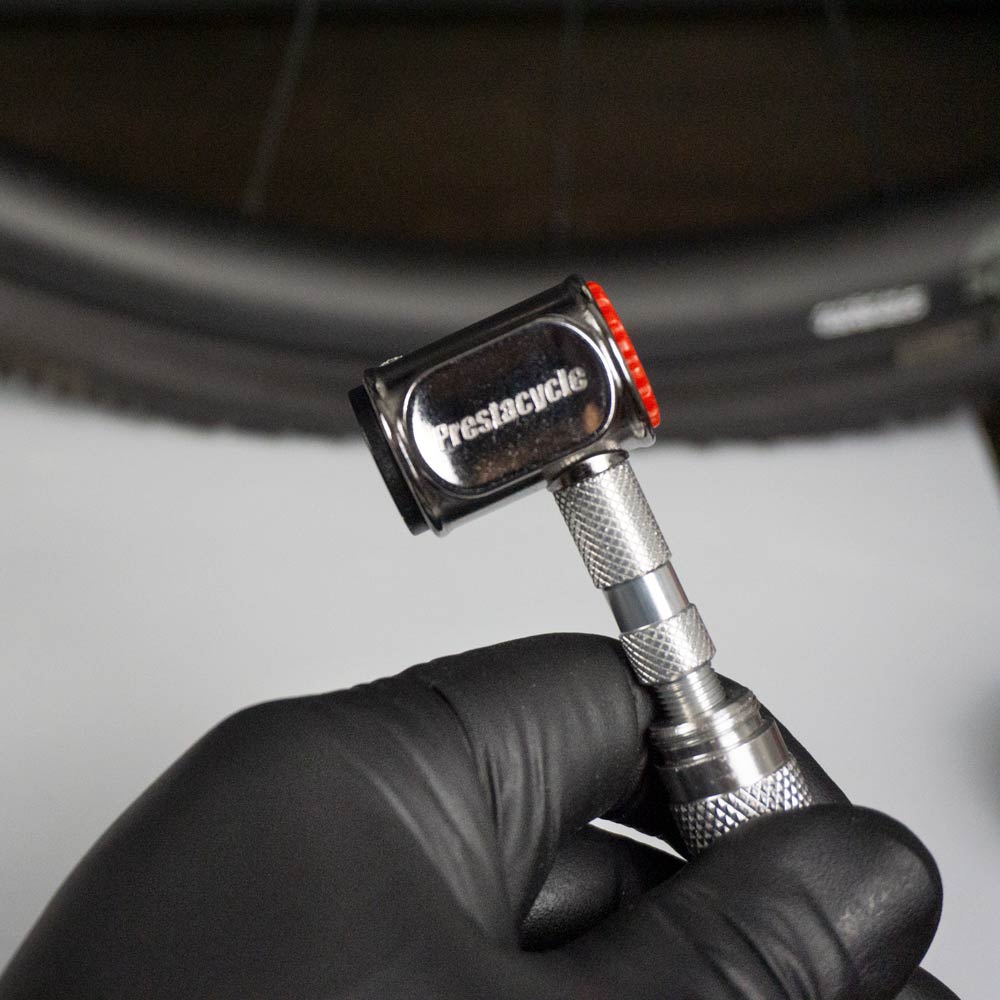 A hand in a black glove holds a Prestacycle Pro Presta Head tool near a bicycle tire, preparing to check or inflate the tire.