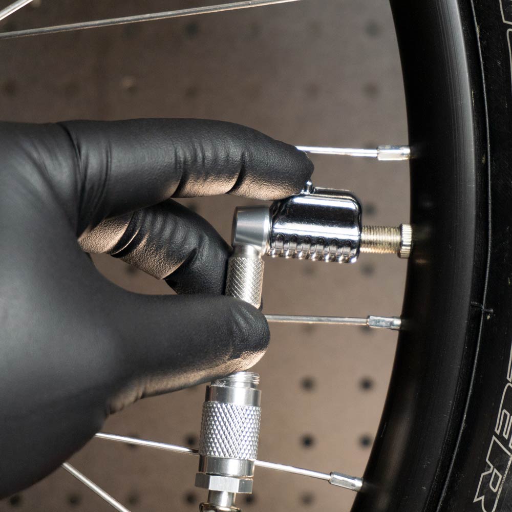 A person in a black glove is attaching the Prestacycle Pro Schrader Head to a bicycle valve stem, preparing to inflate the tire and check pressure. The wheel’s spokes and part of the black rim can be seen.