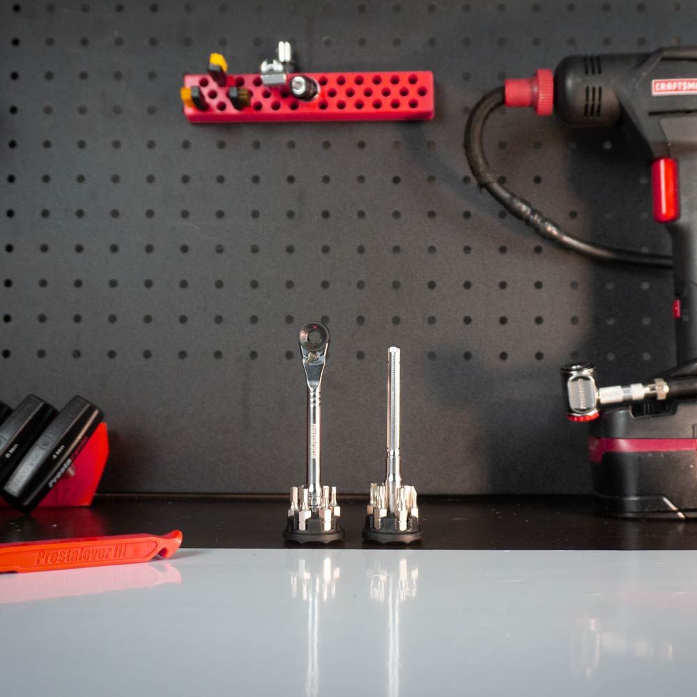 A workbench holds two socket wrenches upright on holders, a red Craftsman power drill, hand tools on the left, a Prestacycle Pro T-Handle Kit with 12 S2 bits and two round holders, plus a red pegboard shelf with small items behind.